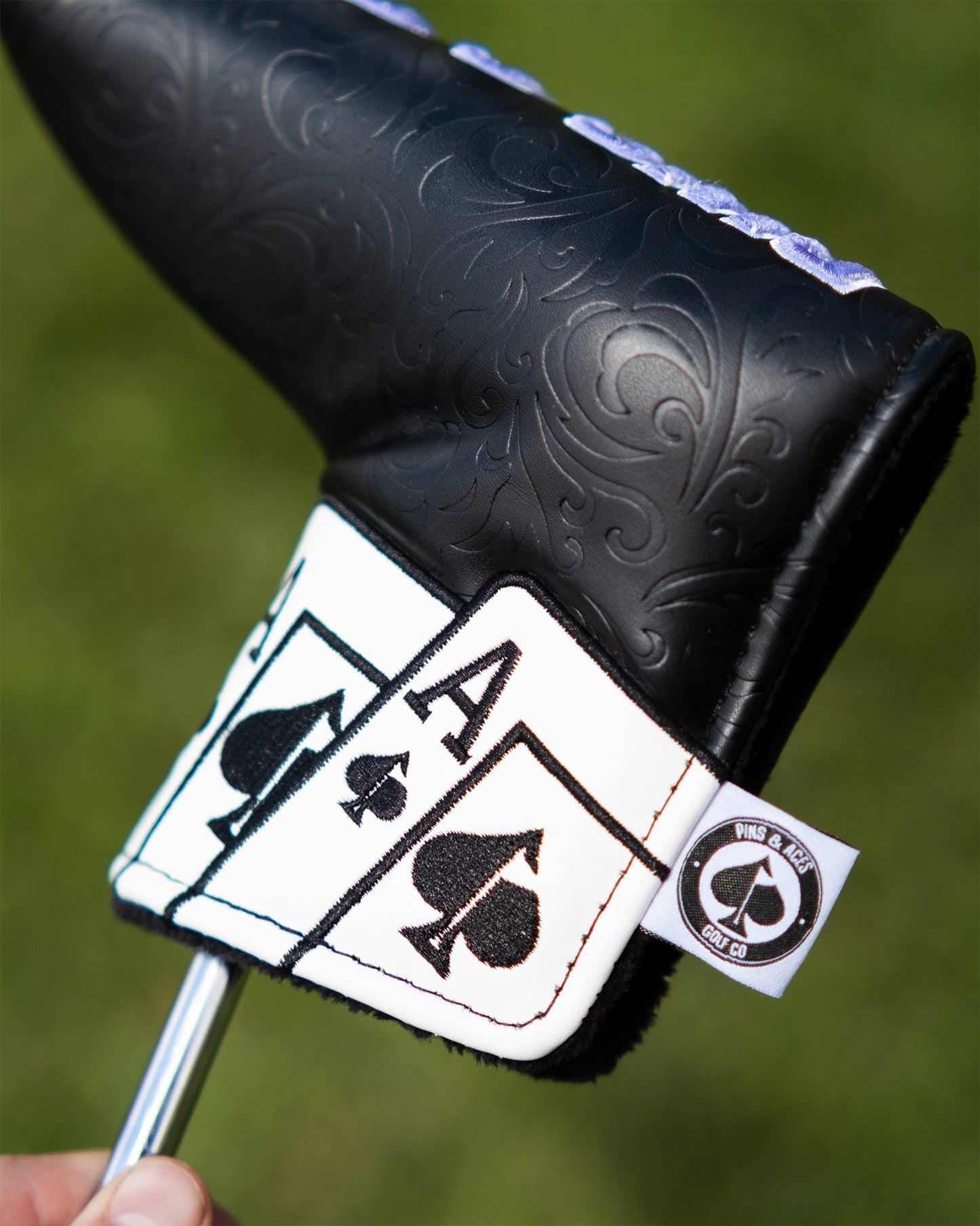 Alt text: Close-up of Ace of Spades blade putter cover with embossed black leather and white details. Features a prominent spade symbol and the ace card design, held in hand against a grassy background. Small logo tag visible on the