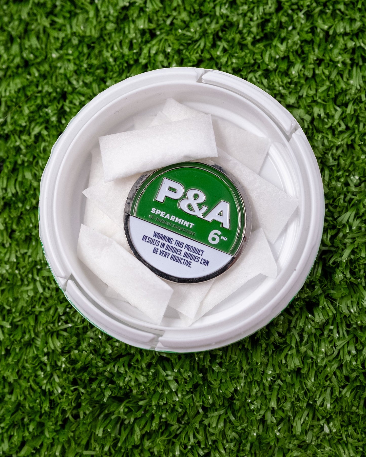 Alt text: "Top view of a 6 Milly Spearmint Ball Marker in a white container with individual pouches, placed on artificial green grass. The container label shows 'P&A, Spearmint, 6 mg'."