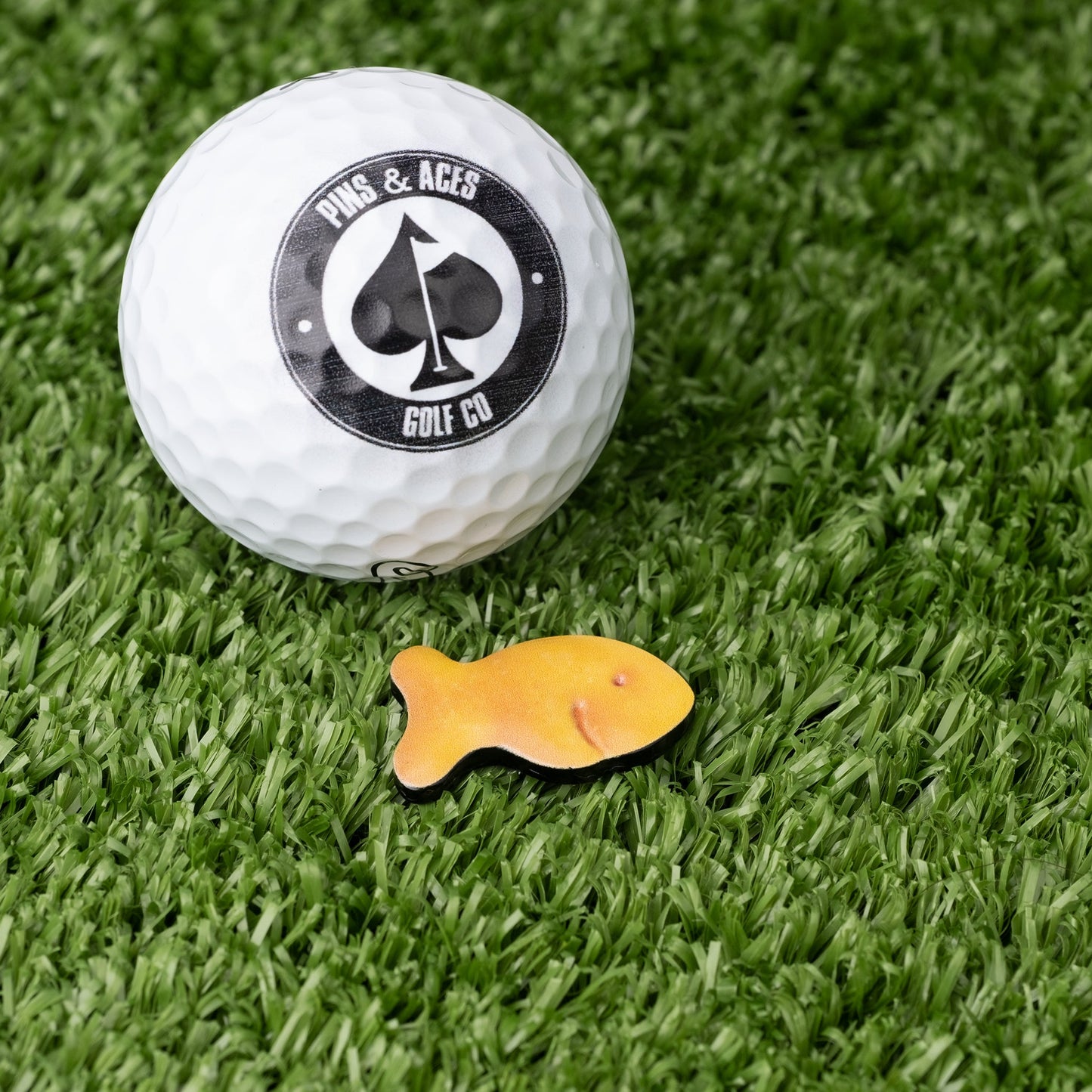 Fish Cracker ball marker next to a golf ball on green turf, adding a playful touch to your golf game.