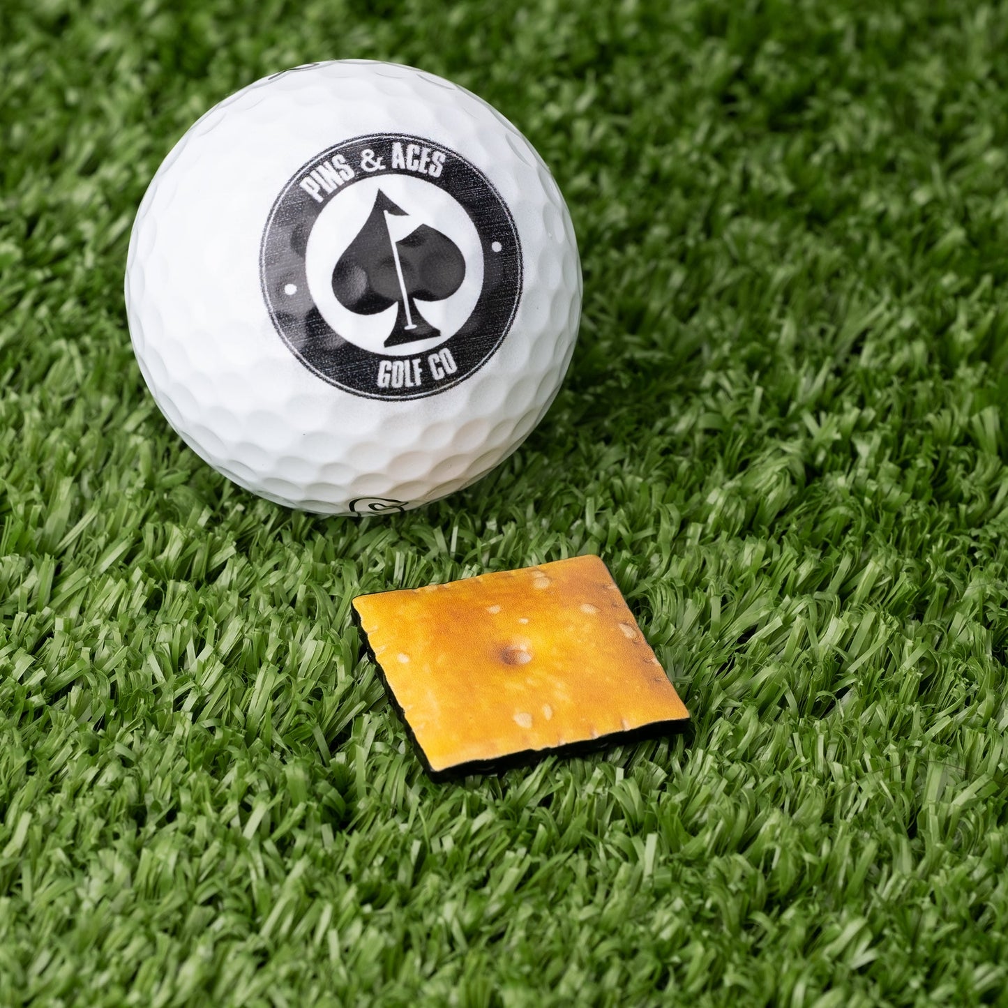 Cheese cracker ball marker next to a golf ball on green turf, showcasing a fun and unique golf accessory.