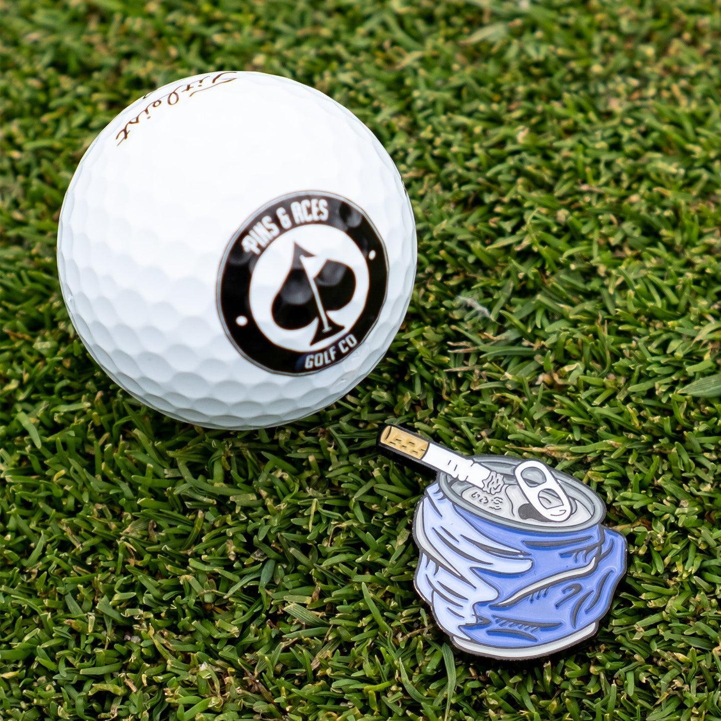 Bold crushed can ball marker beside a golf ball on green grass, perfect for golfers wanting to stand out.