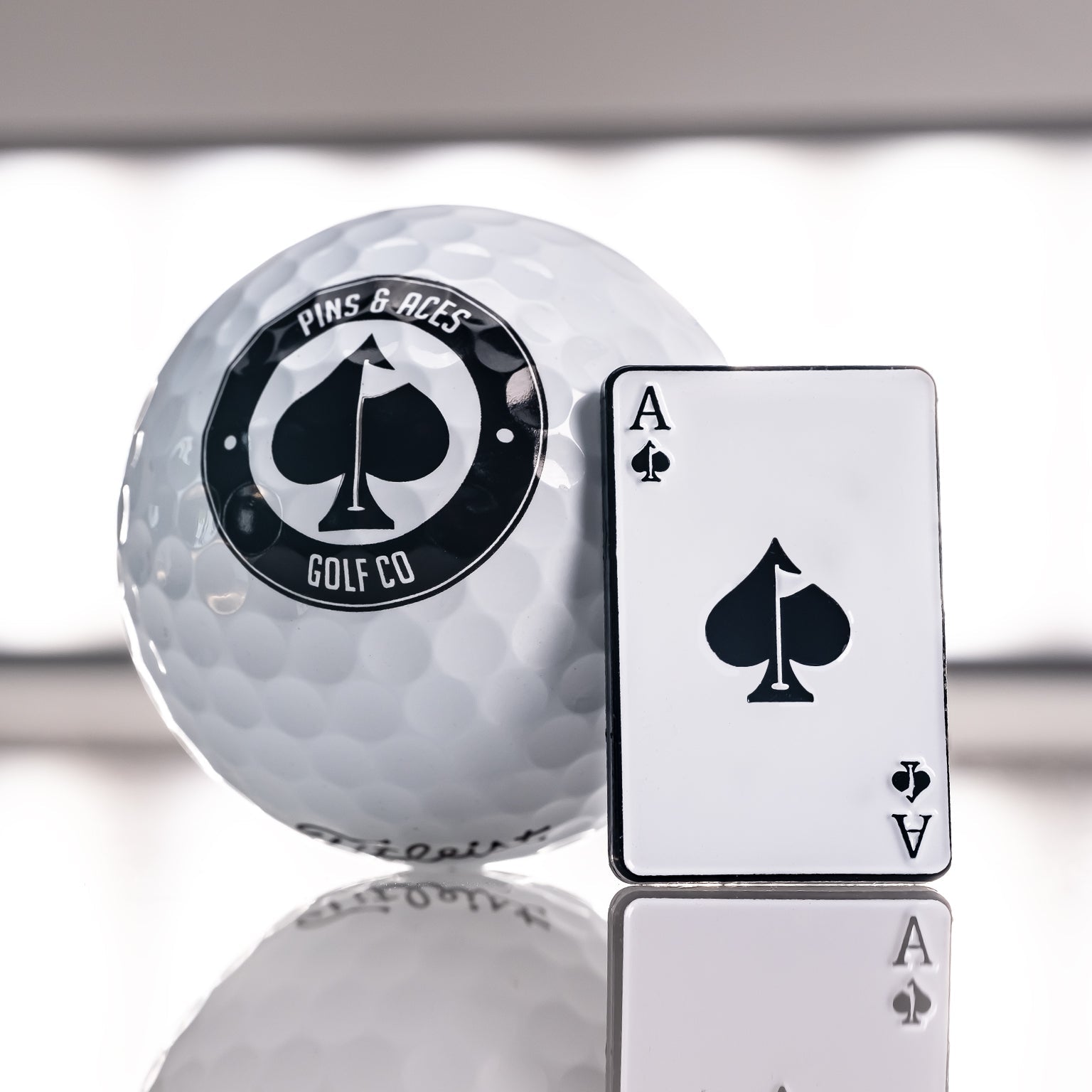 Close-up of an Ace of Spades golf ball marker next to a golf ball with the logo "Pins & Aces Golf Co" on a reflective surface.