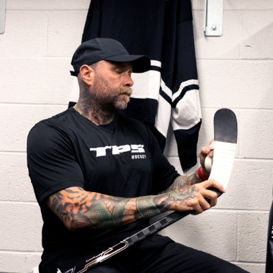 Man sitting in a locker room holding a hockey stick, wearing a black TPS shirt.
