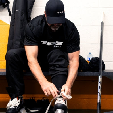 Person tying their shoes in a locker room with hockey equipment.