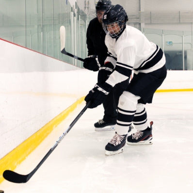 Two ice hockey players on an indoor rink with one player in a white jersey and black helmet, the other in a black jersey and helmet.