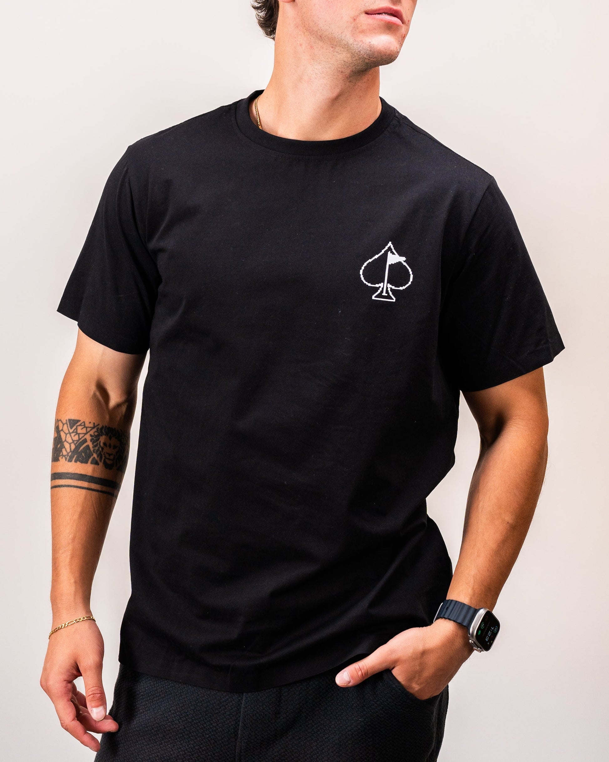 Man wearing a black t-shirt featuring a white spade symbol on the chest, styled with a casual wristwatch and displaying a tattoo on his left arm.