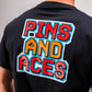 Rear view of a person wearing a black t-shirt with a colorful "PINS AND ACES" text in 8-bit font style printed on the back.