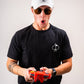 Man in black t-shirt with spade logo playing with a red retro gaming controller, wearing a white cap and sunglasses, looking surprised.