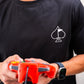 Man wearing a black t-shirt with a white 8-bit spade symbol, holding a red video game controller, and wearing a smartwatch.