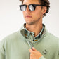 Olive waffle knit pullover with half-zip and logo, worn over a patterned shirt, styled with sunglasses.