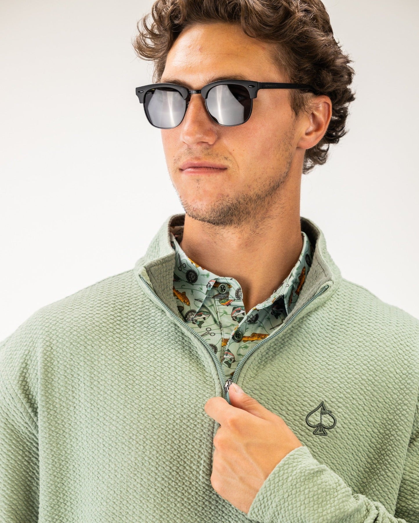 Olive waffle knit pullover with half-zip and logo, worn over a patterned shirt, styled with sunglasses.