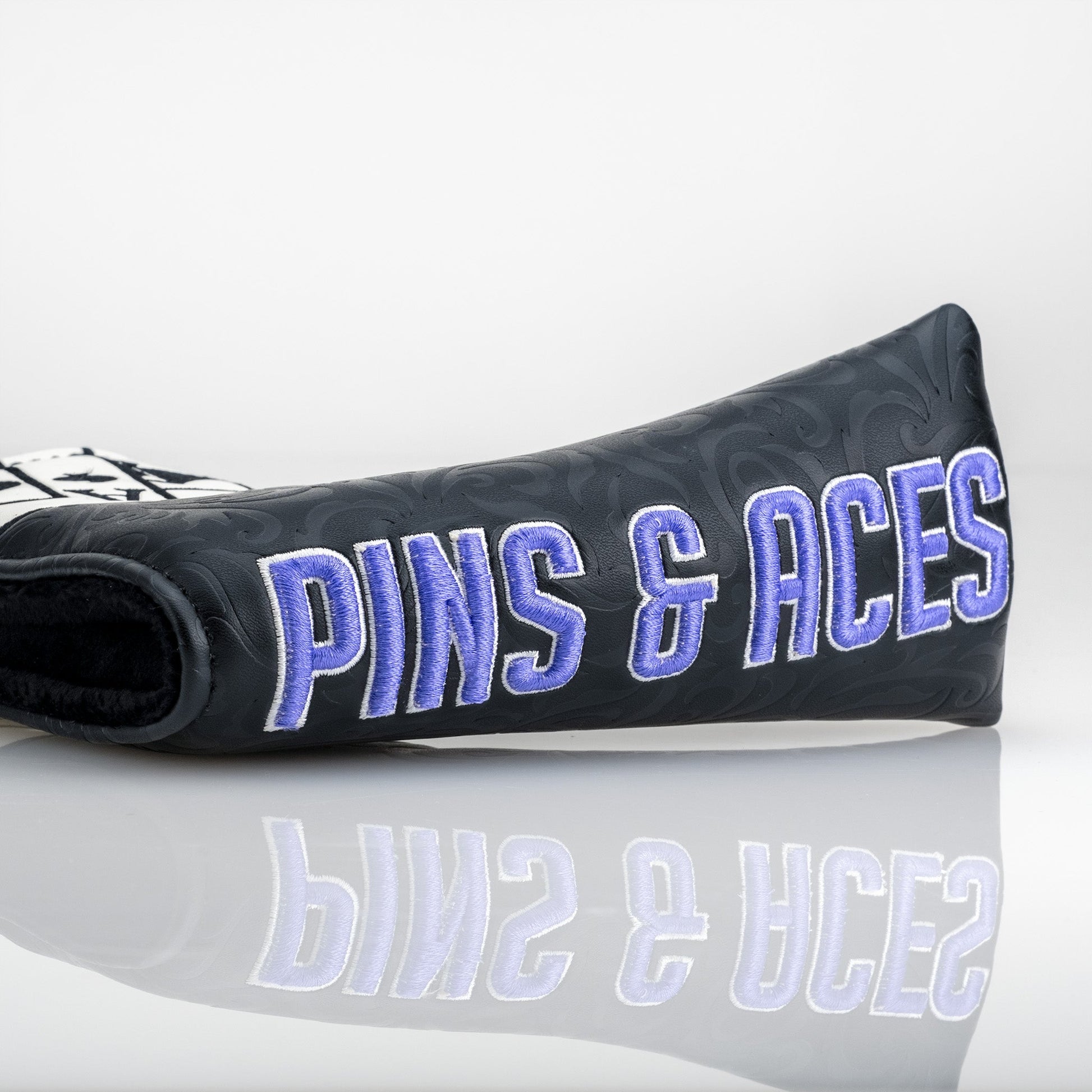 Alt text: "Close-up of a black blade putter cover with embossed patterns, featuring the text 'PINS & ACES' embroidered in blue, displayed against a reflective surface."