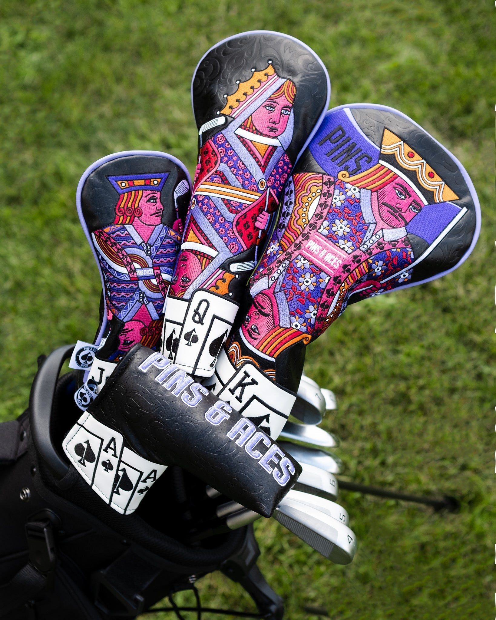 Colorful Ace of Spades blade putter covers in a golf bag on grass, featuring intricate, vibrant designs of playing card royalty in various colors, with the text "Pins & Aces” visible.