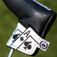 Alt text: Close-up of Ace of Spades blade putter cover with embossed black leather and white details. Features a prominent spade symbol and the ace card design, held in hand against a grassy background. Small logo tag visible on the