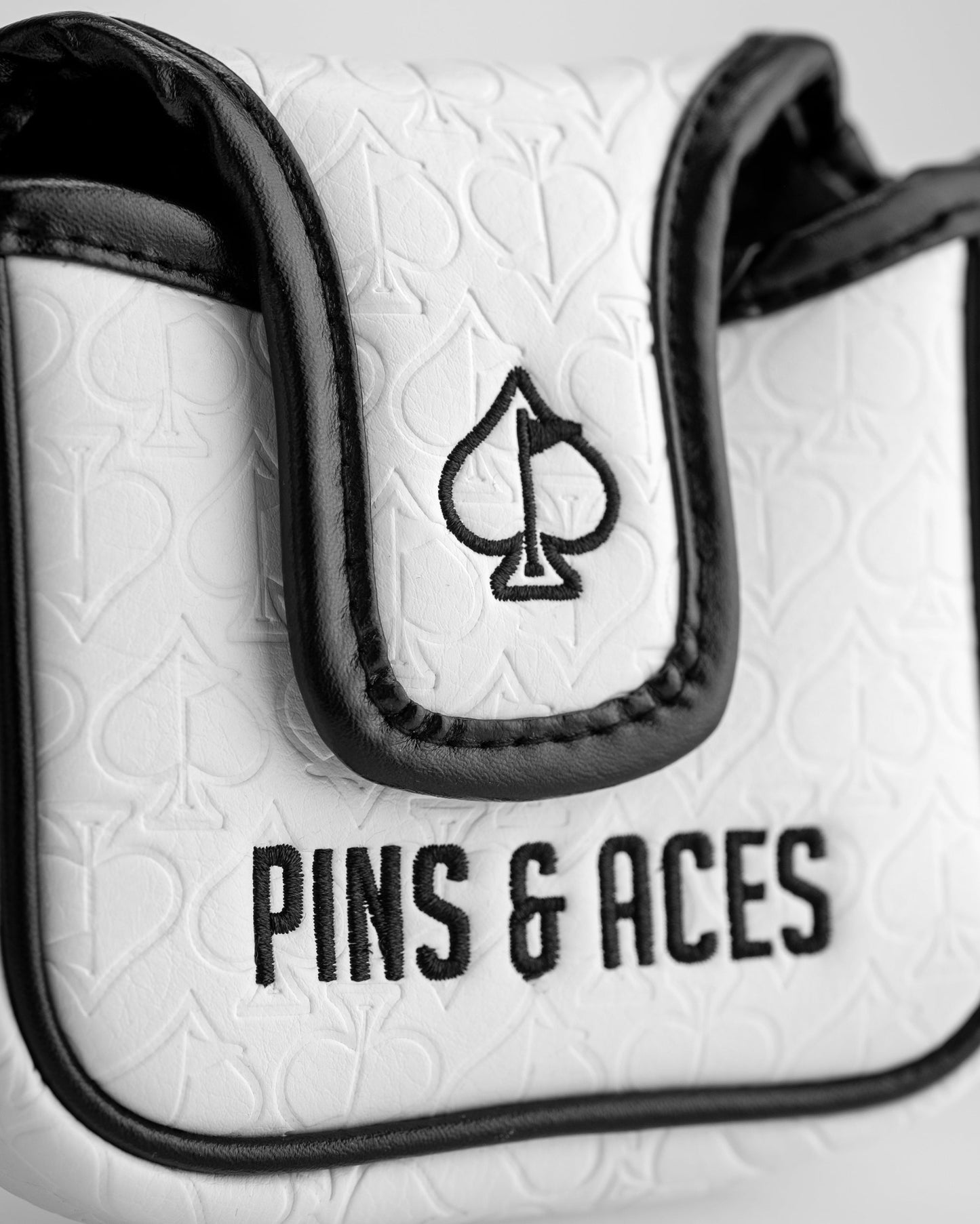 Close-up of a white golf accessory with black trim, featuring a spade logo and "PINS & ACES" embroidery, bold and standout...