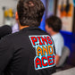 Man wearing a black t-shirt with a colorful "PINS AND ACES" 8-bit retro style graphic on the back, in an arcade setting.