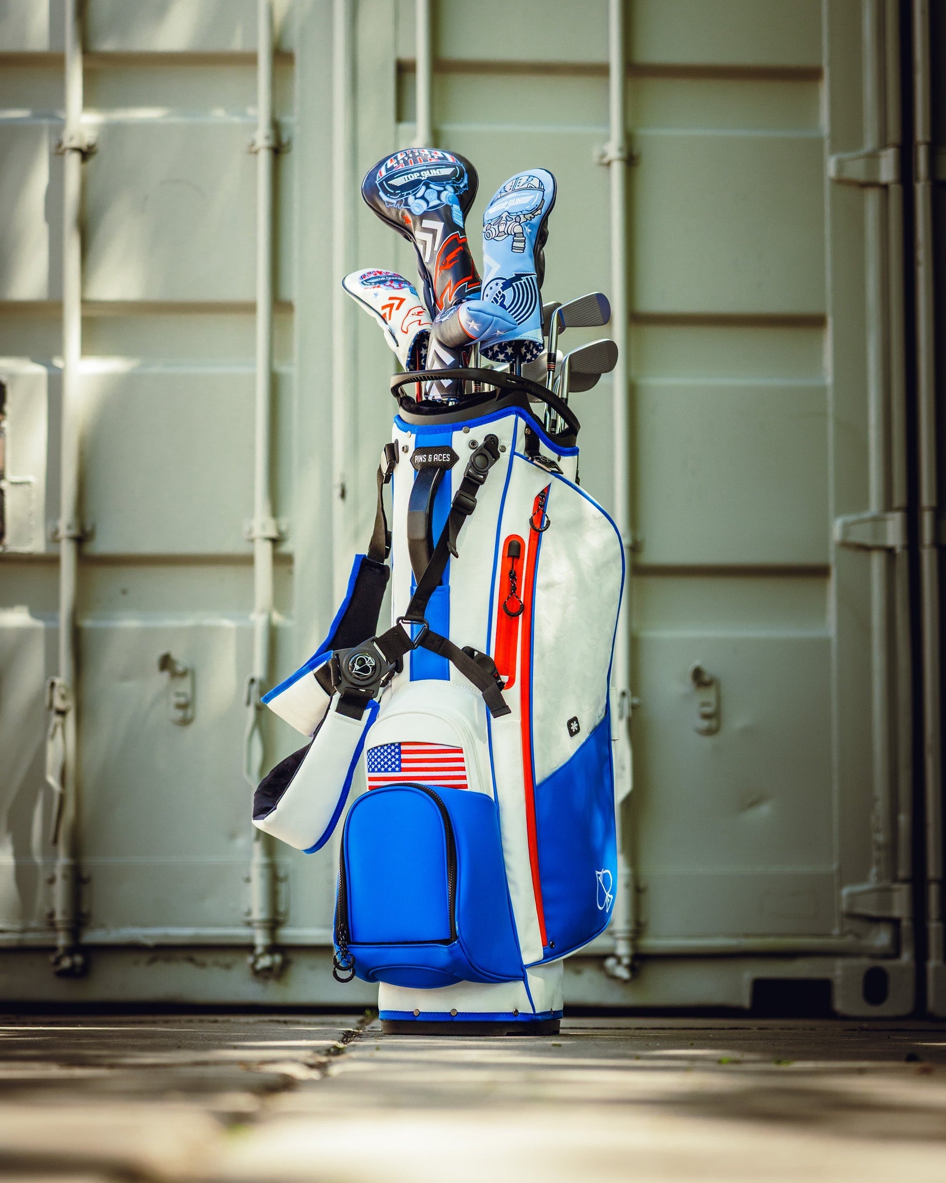 Golf bag with Top Gun Maverick themed blade headcover and golf accessories, standing on a wooden deck near a storage container.