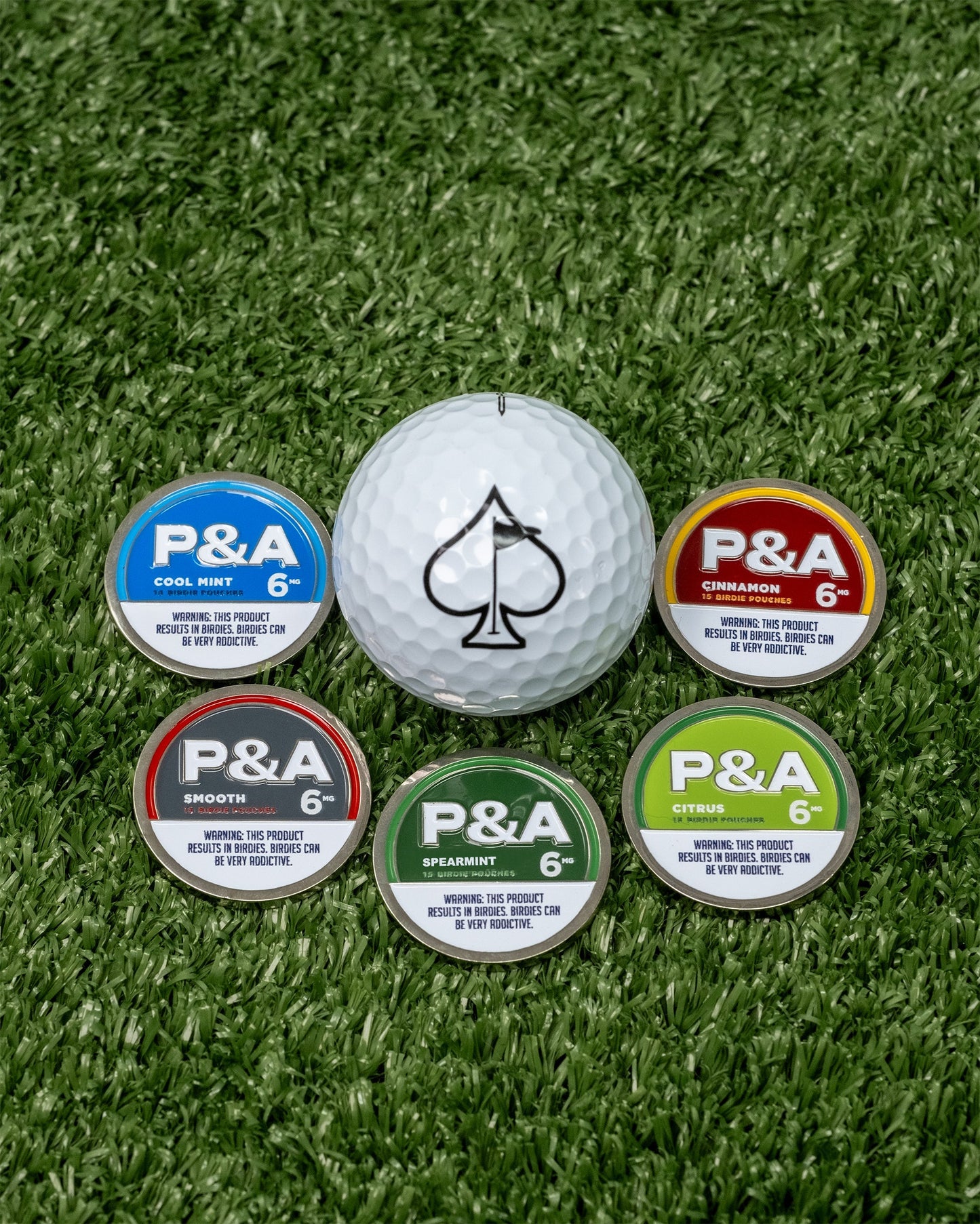 Alt text: "A golf ball with a spade symbol on a dense artificial grass surface, surrounded by five colorful disc-shaped ball markers labeled P&A in flavors Cool Mint, Cinnamon, Smooth, Spearmint, and Citrus."