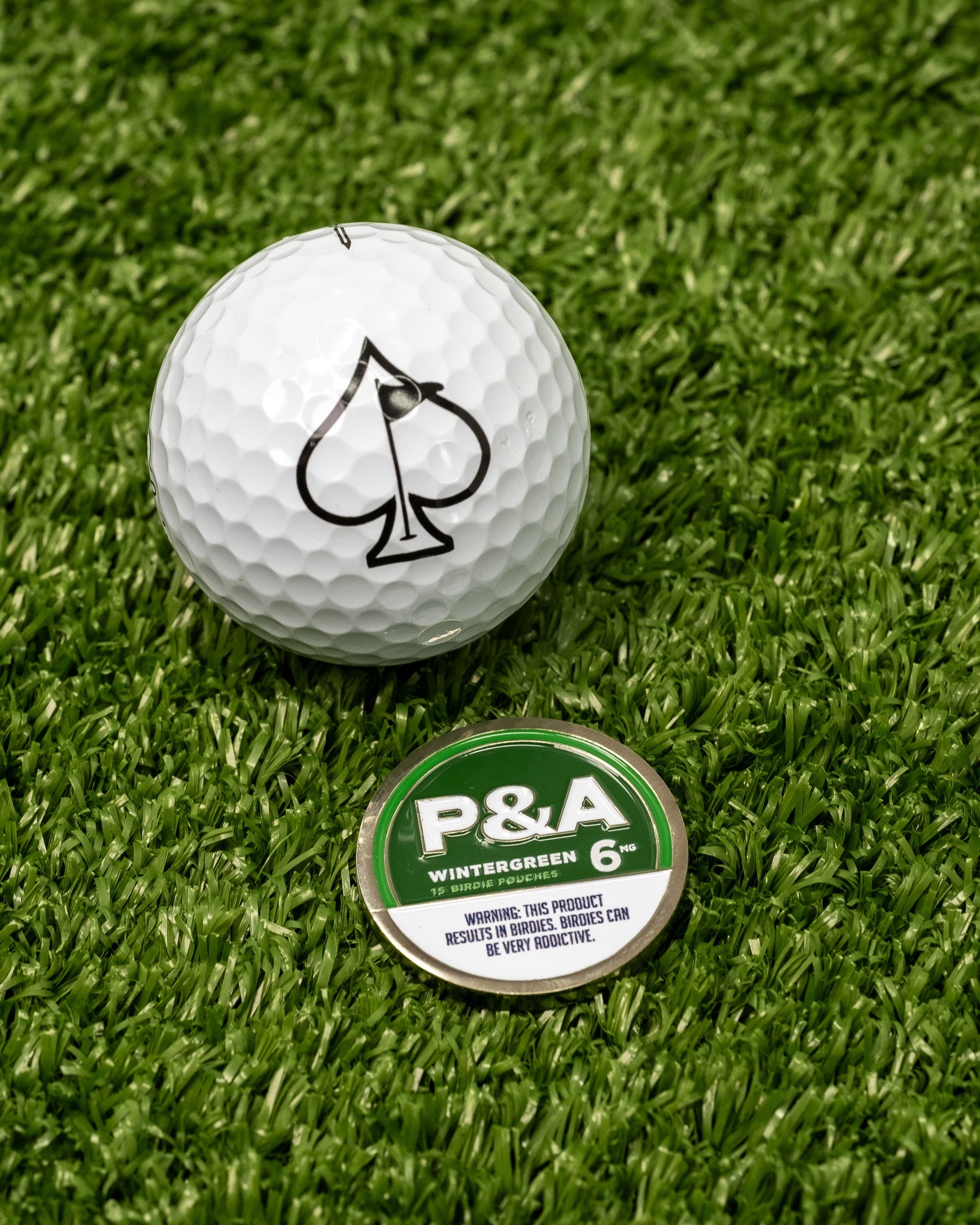 Alt text: "Close-up of a 6 Milly - Wintergreen Ball Marker, featuring a white golf ball with a black spade symbol on artificial grass next to a green and silver Wintergreen 6 marker disc with warning text."
