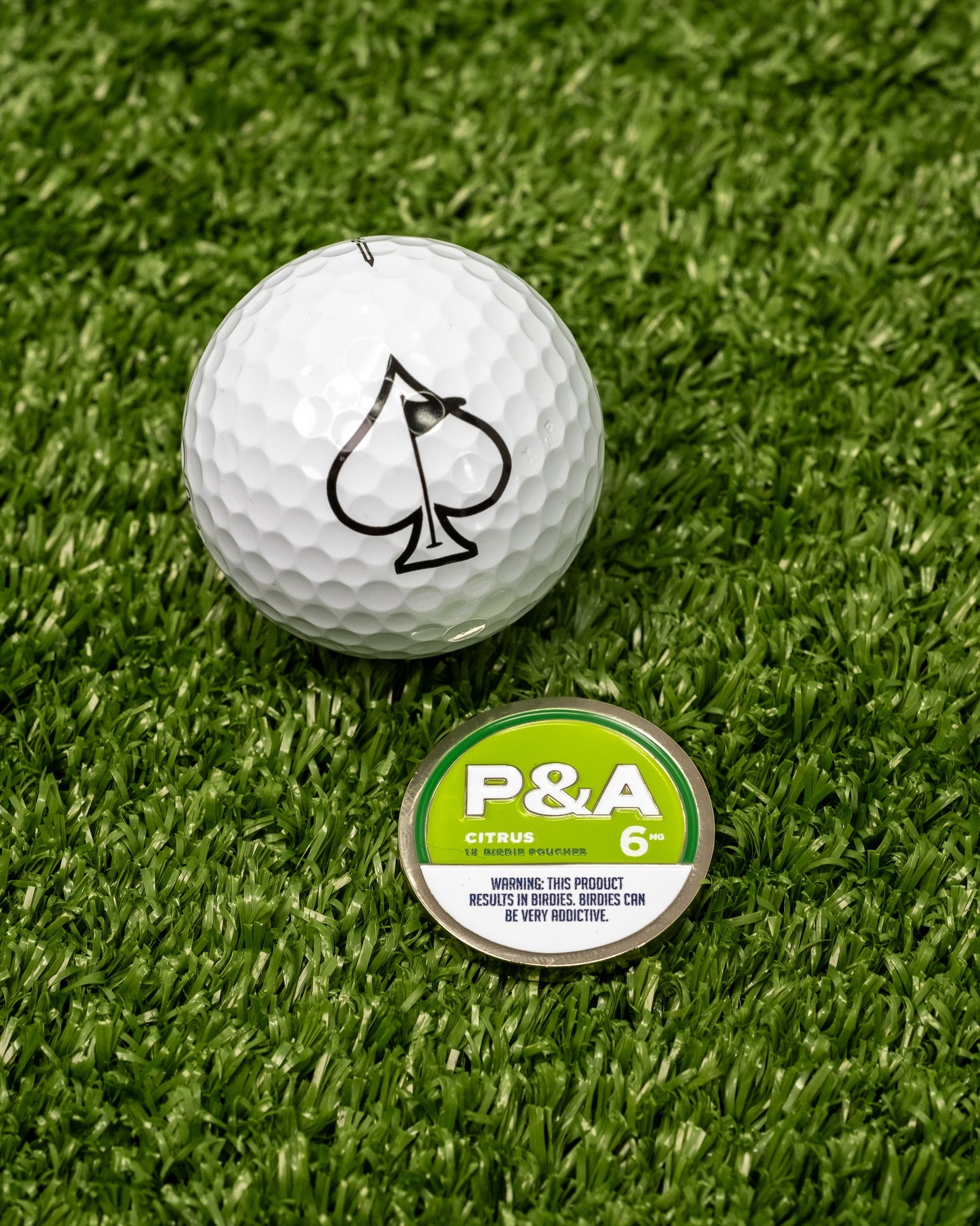 "6 Milly Citrus Ball Marker next to a golf ball with a spade logo, set on green turf.