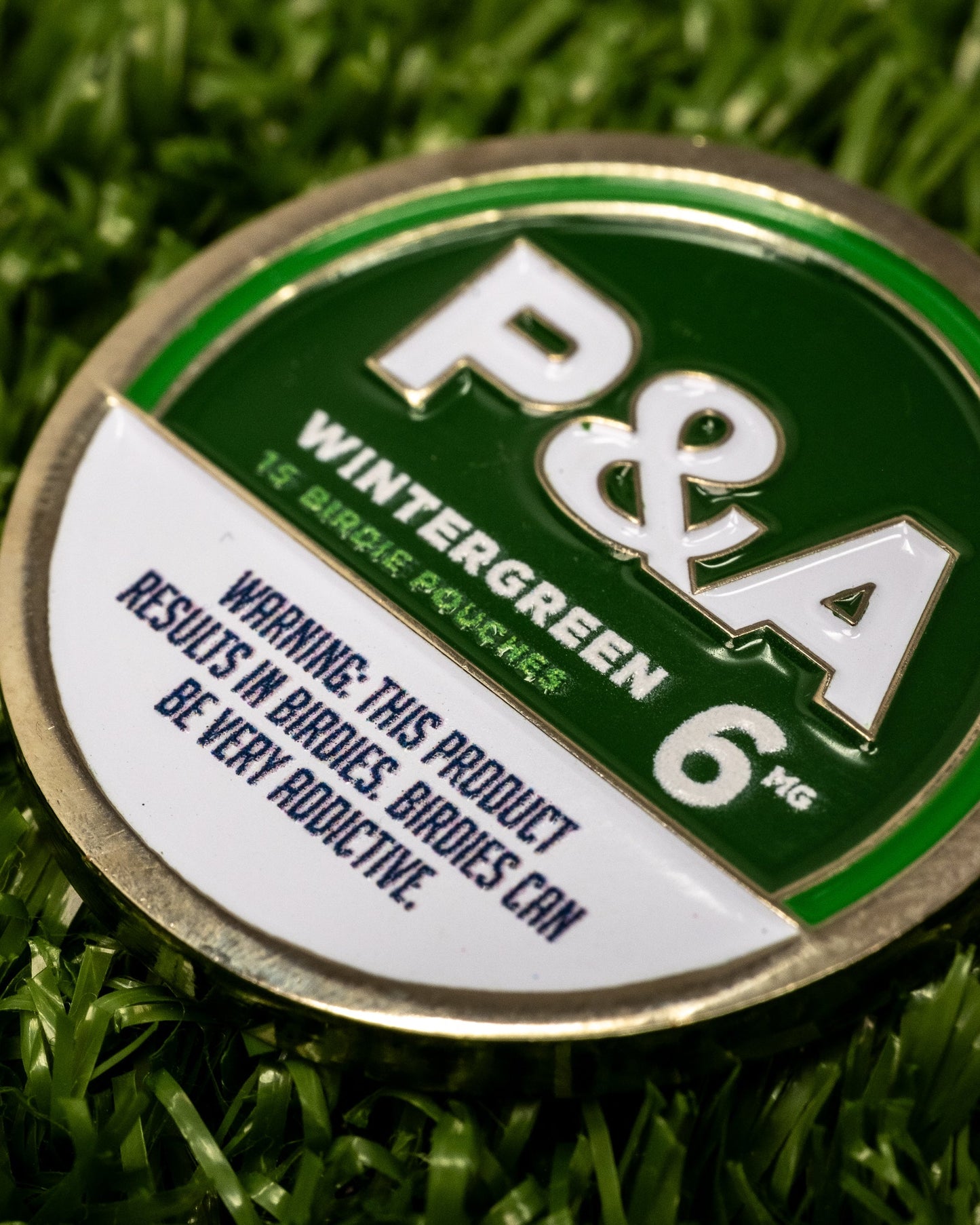Close-up of a 6-milligram Milly Wintergreen golf ball marker placed on artificial grass, featuring a bold green and white color scheme with the text "P&A Wintergreen," and a warning label indicating the product is addictive.