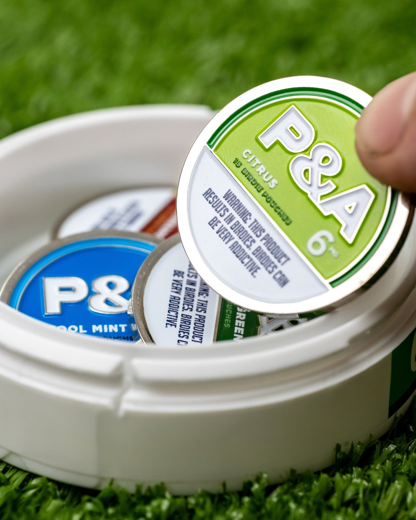 Hand holding a 6 Milly Citrus ball marker from a container with various colored markers on green turf.