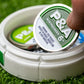Alt text: "Close-up of a hand holding a 6 Milly - Wintergreen ball marker above its storage tin on a background of synthetic grass. The round ball marker features a prominent green and silver design with the logo and the number 6