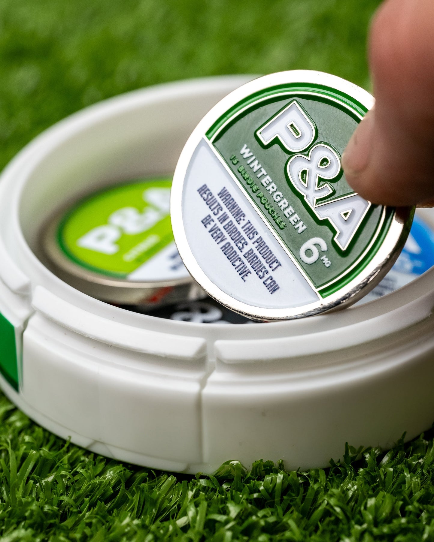 Alt text: "Close-up of a hand holding a 6 Milly - Wintergreen ball marker above its storage tin on a background of synthetic grass. The round ball marker features a prominent green and silver design with the logo and the number 6